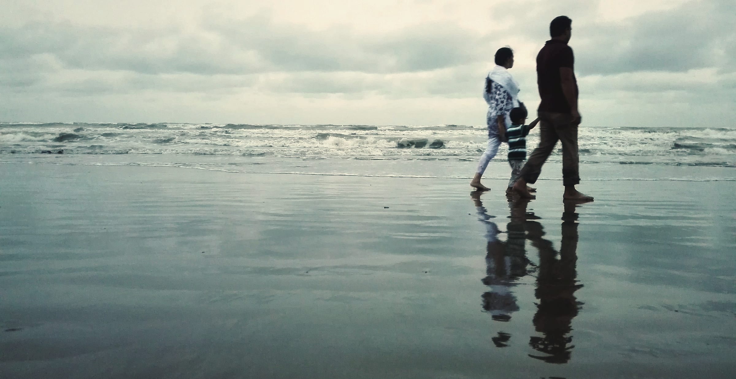Two adults and a child walk hand in hand along a quiet shoreline, their reflections stretching across the wet sand.